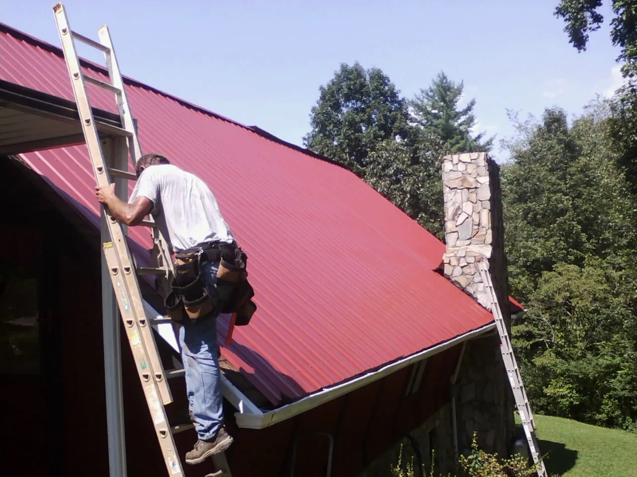 Roofer on a ladder beside a red metal roof and stone chimney — Soffit & Fascia Repair by Roofing Brothers in Carr